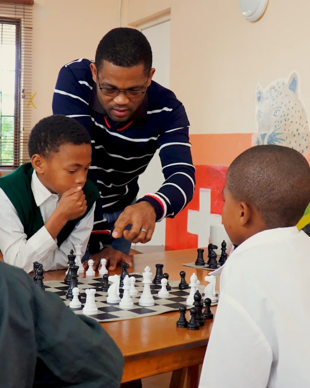 A coach leaning over the table to point out a move while two boys study the chess board