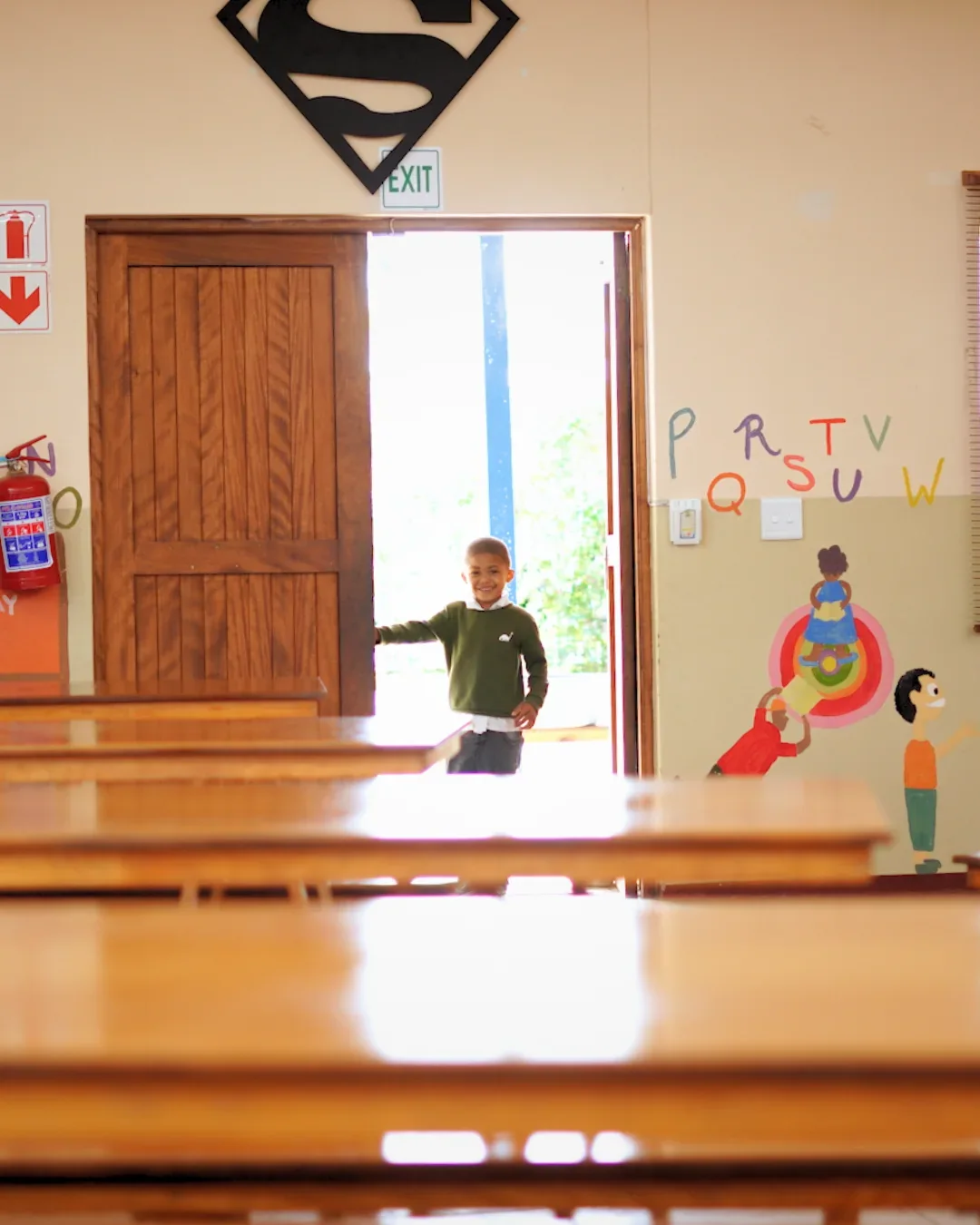 A boy in a green jersey standing in the open doorway of the classroom, desks in the foreground