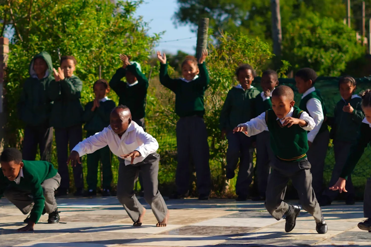 Children racing across the paved yard in school uniform while the rest of the group cheers them on