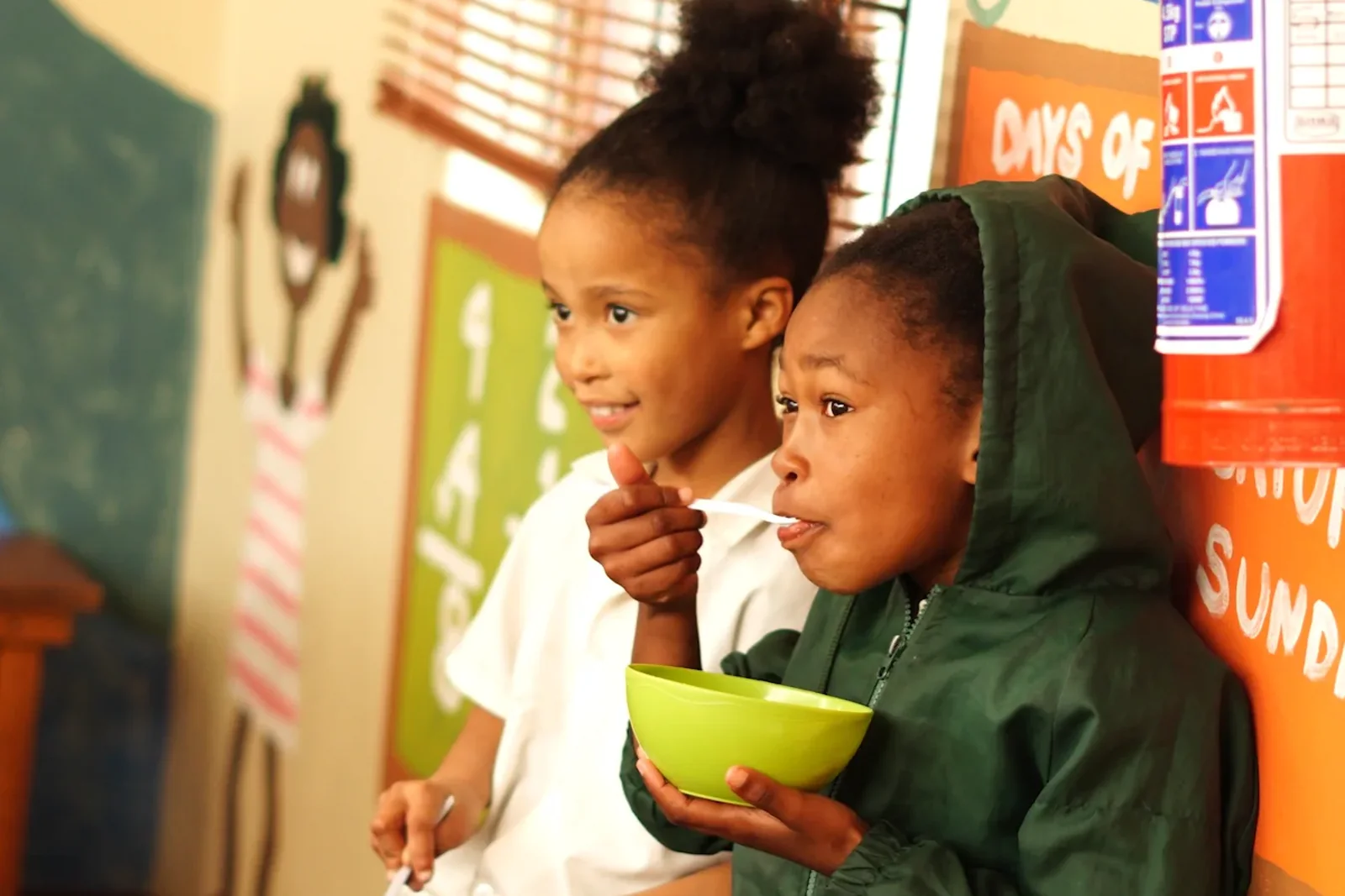 Two children sitting against the classroom wall eating from bowls, one in a green jacket