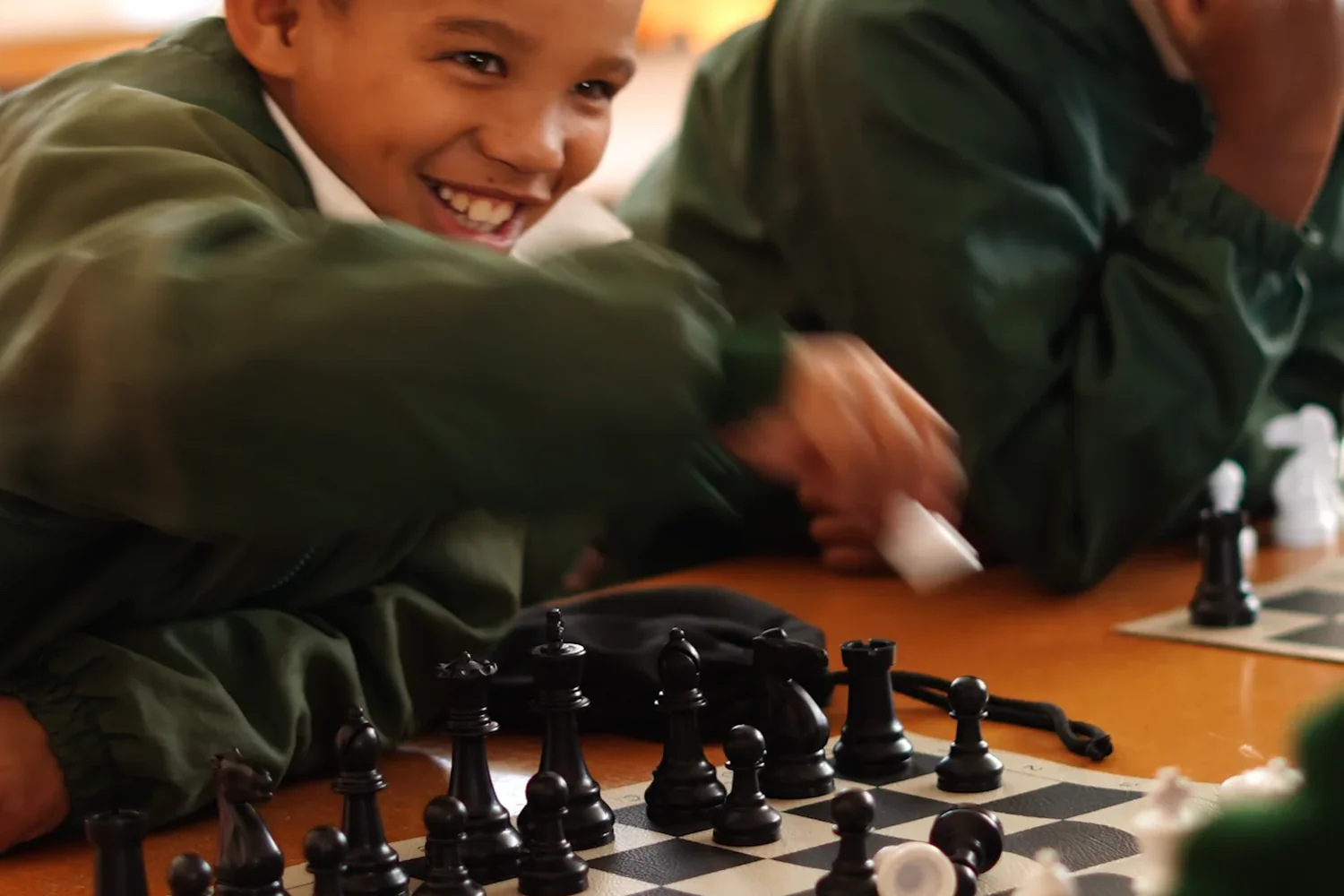 A boy grinning over his pieces mid-game, another player thinking at the board beside him