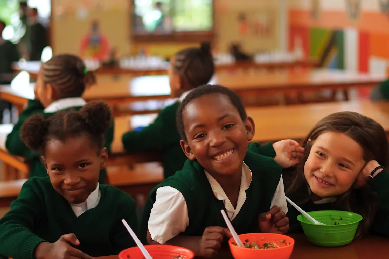 Three children at the feeding table turning to smile, bowls and spoons in front of them