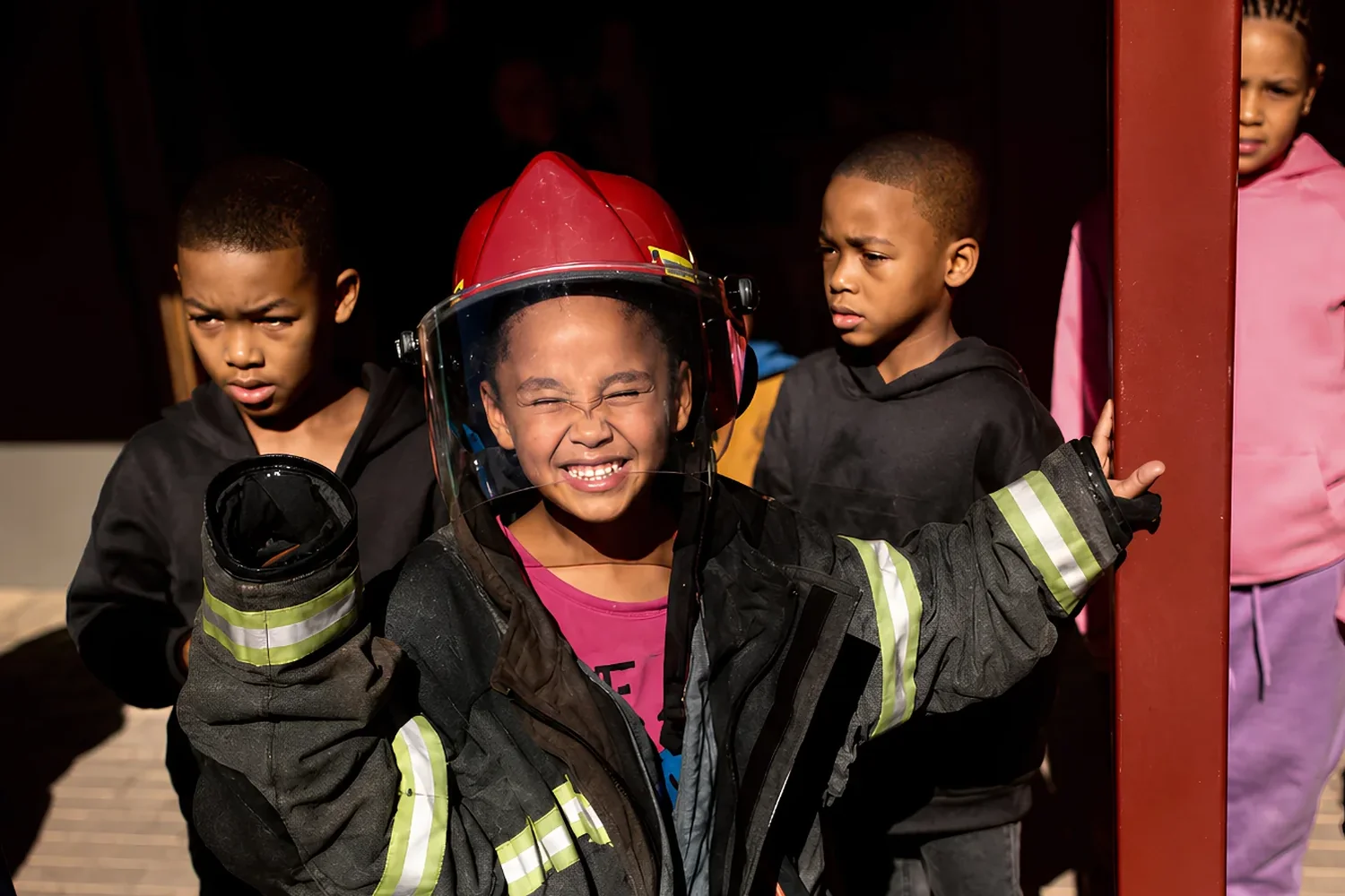 A girl grinning in an oversized firefighter jacket and helmet, other children behind her
