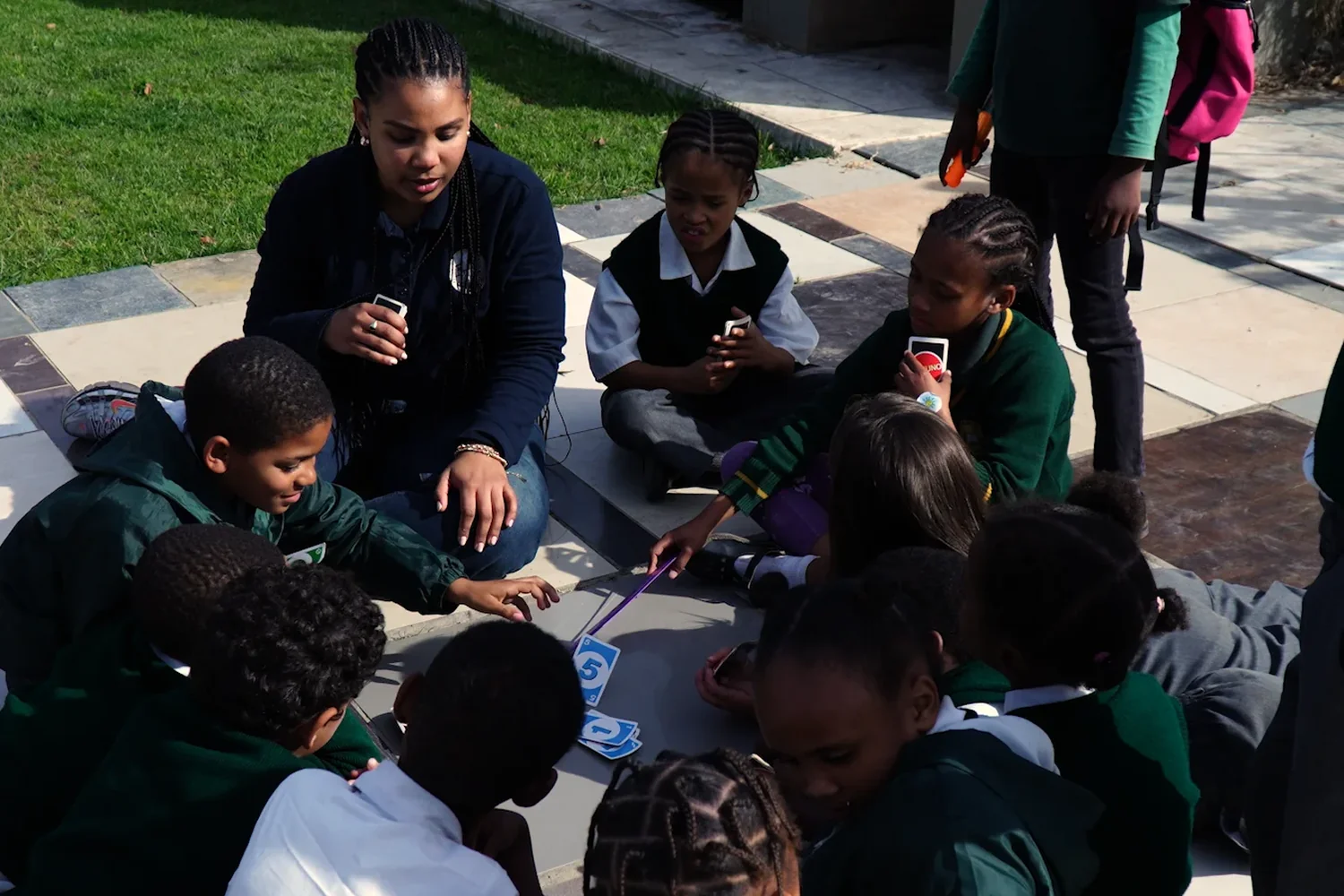 An older girl sitting on the paving leading a card game with a circle of younger children