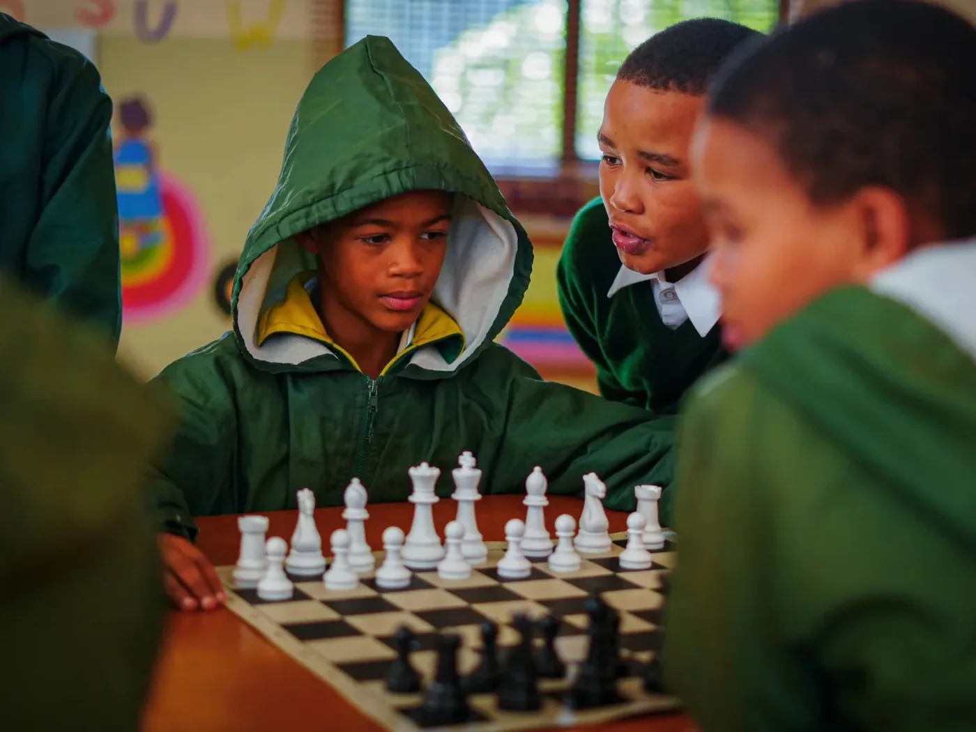 Children around a chess board in the classroom, one leaning in to talk through the position with the player