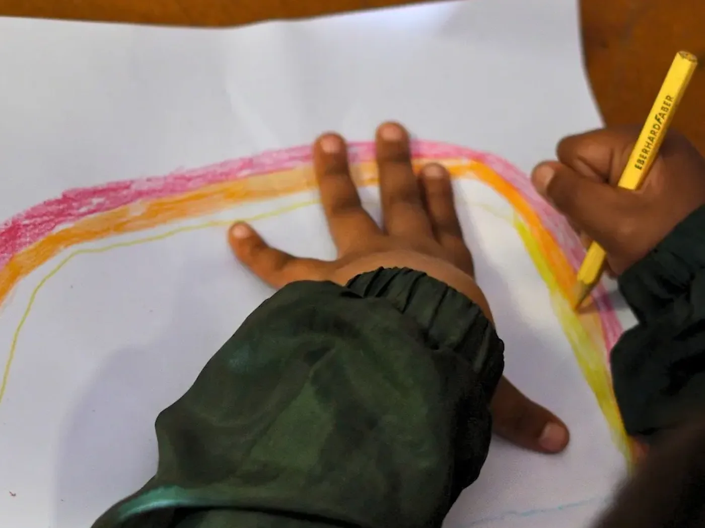 A child's hand resting on a sheet of paper, drawing a rainbow in coloured pencil, with a tub of pencils beside it